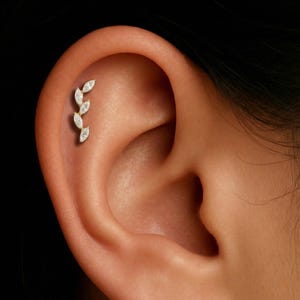 May include: A gold-colored helix earring featuring a leaf design with four clear, marquise-cut stones. The earring is positioned on the upper ear cartilage, showcasing a delicate and elegant aesthetic.