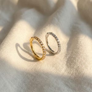 Peut inclure: Deux boucles d'oreilles créoles, une dorée et une argentée, chacune incrustée de petits cristaux transparents. Les boucles d'oreilles sont présentées sur une surface blanche texturée, avec un éclairage doux créant des ombres.