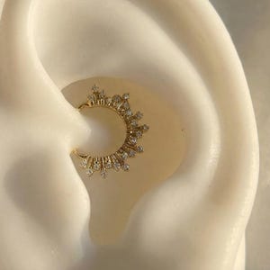 Può includere: Un orecchino dorato con un design a raggiera, ornato da piccoli cristalli scintillanti. Il gioiello è posto su uno sfondo bianco sporco, che mette in risalto i dettagli intricati dell'accessorio per l'orecchio.