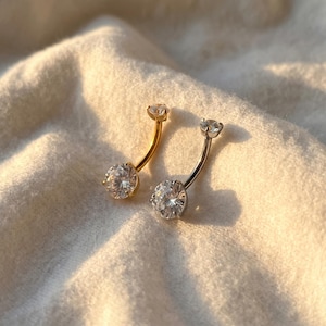 May include: Two belly button rings. One is gold-toned, the other is silver-toned. Each has a curved barbell design with a large, clear, round gemstone at the bottom and a smaller gemstone at the top. The jewelry is displayed on a soft, white fabric.