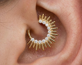 May include: Gold-coloured ear piercing jewellery with a sunburst design. The jewellery features a series of small, sparkling clear stones set in a curved shape, with radiating spikes. The jewellery is worn in the ear.