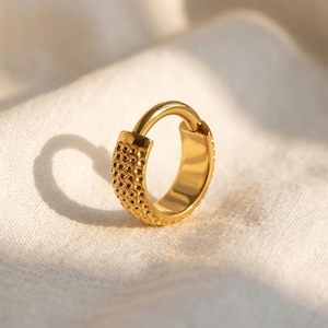 May include: A gold-colored hoop earring with a textured, dotted design on the front. The earring is a single piece with a hinged closure. The earring is set against a white, textured background.
