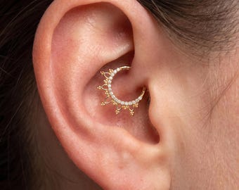 May include: A gold hoop earring featuring a semi-circular row of small clear stones and a decorative outer edge with pointed accents. The jewellery is positioned within the inner cartilage of an ear, showcasing a hinged closure mechanism.