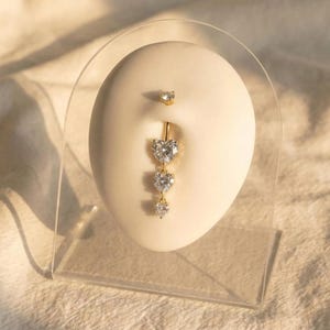 Könnte beinhalten: Ein goldfarbener Bauchnabelring mit einem hängenden Herzdesign. Der Ring hat einen kleinen Edelstein oben und drei herzförmige Edelsteine, die darunter hängen. Der Schmuck wird auf einer cremefarbenen Form präsentiert.