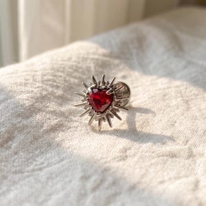 May include: A silver-toned metal stud earring featuring a central heart-shaped red gemstone. The stone is surrounded by a sunburst-style frame with multiple sharp, radiating spikes extending outward from the centre, creating a star-like appearance.
