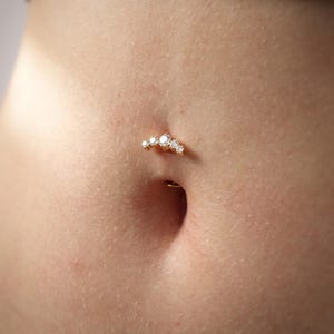 May include: A gold navel piercing jewelry piece featuring a curved bar design adorned with five clear, round-cut stones arranged in a graduated arc. The jewelry is positioned within the navel, showcasing a metallic gold finish and sparkling stones.