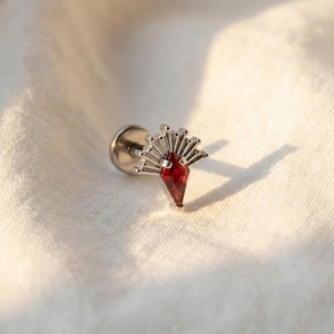 May include: A silver stud earring featuring a fan-shaped design and a red gemstone. The earring has a post and a flat back. The gemstone is a deep red colour and is cut into a diamond shape.