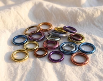May include: A selection of colourful metal rings in various sizes and shades, including gold, blue, purple, and bronze. The rings are scattered on a white, textured surface, creating a visually appealing arrangement.