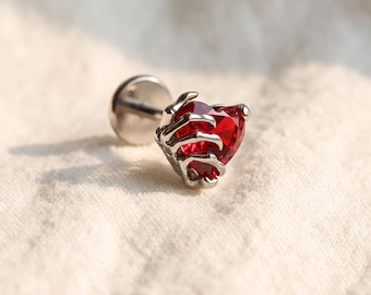 May include: A silver-toned metal stud earring featuring a skeletal hand design clutching a faceted, heart-shaped red gemstone. The piece includes a flat circular backing post, showcasing a detailed metallic claw grip around the red heart.