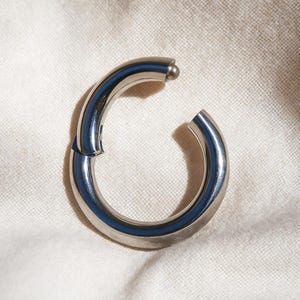 May include: A polished, silver-toned hinged hoop earring with a small ball closure. The earring features a blue stripe on the inside and is open at one end. It rests on a white, textured surface.