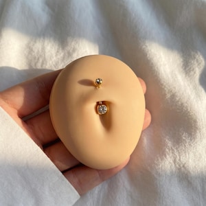 May include: Gold-colored belly button ring with a small, clear gem at the bottom and a smaller gem at the top. The jewelry is displayed on a tan-colored, oval-shaped form, simulating a belly. The background is a white, textured fabric.