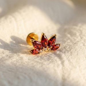 Op de afbeelding: Een gouden oorbel met een bloemmotief. Het heeft rode marquise-geslepen edelstenen en kleine witte edelstenen. De oorbel is geplaatst tegen een witte, getextureerde achtergrond.