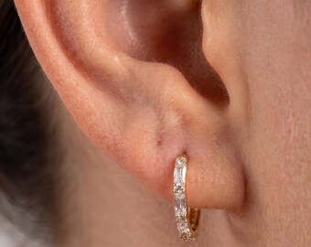 May include: A small gold hoop earring featuring a series of clear, rectangular baguette-cut stones set along the front edge. The earring is secured through the earlobe, showcasing a polished metallic finish and sparkling, faceted gems.