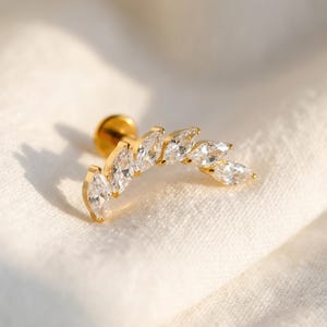 May include: A gold-colored ear stud featuring a curved design of six clear, marquise-cut cubic zirconia stones. The earring is set on a gold post and backing, resting on a white fabric surface. The overall design is elegant and suitable for various occasions.