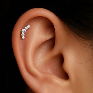 May include: A close-up of an ear with a curved, silver-toned earring. The earring features a series of graduated, clear, round-cut gemstones. The gemstones are set closely together, creating a sparkling, elegant design.