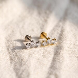 May include: Two flat back stud earrings, one in silver tone and one in gold tone, each featuring a trio of clear round prong-set cubic zirconia stones arranged in a horizontal row. The studs have a circular base and a straight post for insertion.