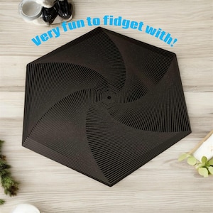 Puede incluir: Juguete fidget hexagonal negro con diseño en espiral. El texto "Very fun to fidget with!" está impreso en azul. El juguete está sobre una superficie de madera clara.