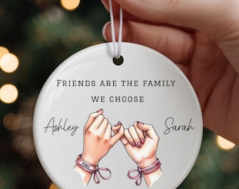 Puede incluir: Adorno navideño de cerámica blanca con el texto "FRIENDS ARE THE FAMILY WE CHOOSE". Presenta ilustraciones de dos manos haciendo una promesa de meñique, con los nombres "Ashley" y "Sarah" y el año "2025".