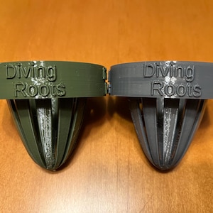 May include: Two 3D-printed objects, one olive green and one gray, with the text "Diving Roots" on the top. The objects have a cylindrical top and a cone-shaped, slotted bottom. The background is a light brown wooden surface.