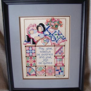 May include: Framed cross-stitch artwork featuring two figures sitting on a quilt with the text "May your sorrows be patched and your joys be quilted".
