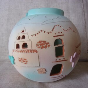 May include: A round, off-white ceramic vase with a light blue top and base. The vase features a Southwestern design with cut-out windows and architectural details in shades of orange and pink. Decorative cacti and clouds are also included.