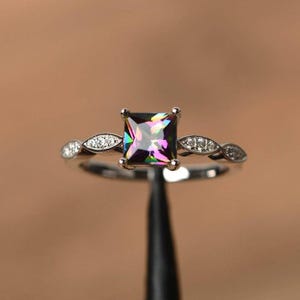 Könnte beinhalten: Ein silberner Ring mit einem quadratischen, mehrfarbigen Edelstein als Mittelpunkt. Das Band hat ein zartes Design mit kleinen, klaren Steinen, die in einem blattartigen Muster auf beiden Seiten des Hauptsteins angeordnet sind. Der Ring wird vor einem verschwommenen Hintergrund gezeigt.