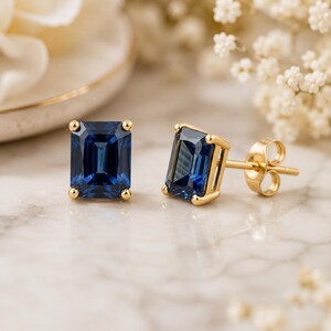 Könnte beinhalten: Ein Paar Ohrstecker mit smaragdgeschliffenen, tiefblauen Edelsteinen in Gelbgoldfassungen. Ein Ohrring ist von vorne zu sehen, während der andere im Seitenprofil positioniert ist und den Goldstift sowie den Schmetterlingsverschluss zeigt.