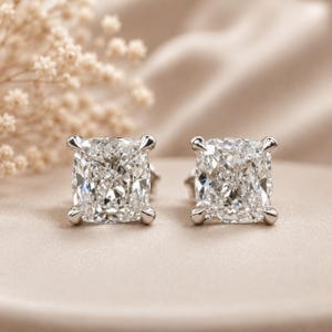 May include: A pair of stud earrings featuring cushion-cut clear gemstones set in four-prong silver-toned metal settings. The stones are faceted to reflect light, showcasing a brilliant white appearance. The earrings are positioned side by side.
