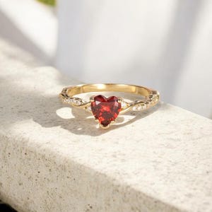 May include: A gold ring with a heart-shaped, red gemstone. The band features a twisted design with small, clear accent stones. The ring is set against a neutral background, highlighting its intricate details and vibrant color.