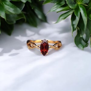 May include: A gold ring with a marquise-cut, deep red gemstone. The ring's band features an intricate, braided design with floral accents. The ring is set against a white background with green foliage in the background.
