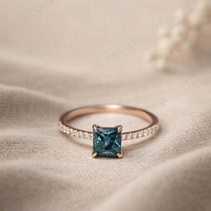 Może przedstawiać: Pierścionek z różowego złota z kwadratowym, turkusowym kamieniem szlachetnym pośrodku. Obręcz jest inkrustowana małymi, przezroczystymi kamieniami szlachetnymi. Pierścionek jest umieszczony na neutralnym, teksturowanym tle.