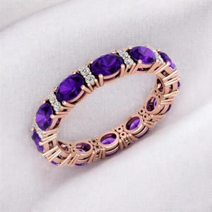 Może przedstawiać: Pierścionek z różowego złota z owalnymi fioletowymi kamieniami szlachetnymi i małymi białymi akcentami. Pierścionek jest osadzony na białym tle, eksponując wzór i kolory kamieni szlachetnych. Biżuteria.