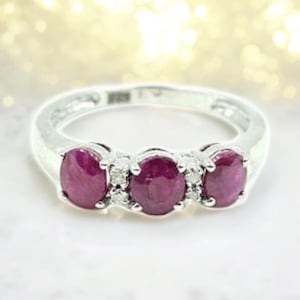 Może przedstawiać: Srebrny pierścionek z trzema owalnymi, głęboko czerwonymi kamieniami szlachetnymi, prawdopodobnie rubinami. Małe, przezroczyste kamienie akcentujące są wstawione między większymi kamieniami. Pierścionek ma wypolerowaną, odbijającą światło powierzchnię.