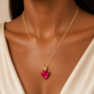 Puede incluir: Un collar de cadena dorada con un colgante de piedra preciosa roja en forma de corazón facetado. El colgante está asegurado en un engaste dorado y cuelga de una delicada cadena de eslabones dorados que descansa sobre la zona de la clavícula.