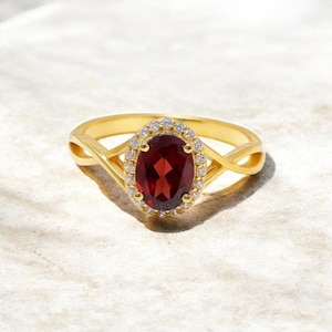 Puede incluir: Un anillo de oro con una piedra preciosa ovalada de color rojo intenso, rodeada por un halo de piedras más pequeñas y claras. La banda del anillo tiene un diseño intrincado e entrelazado. El anillo está sobre un fondo neutro y texturizado.