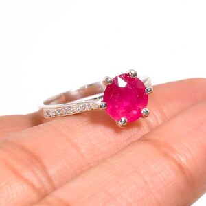 Może przedstawiać: Srebrny pierścionek z dużym, okrągłym, jasnoróżowym kamieniem szlachetnym. Obręcz pierścionka zdobią mniejsze, przezroczyste kamienie. Pierścionek jest prezentowany na dłoni, podkreślając wzór i kolor biżuterii.