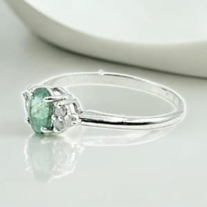 Może przedstawiać: Srebrny pierścionek z jasnozielonym owalnym kamieniem szlachetnym. Kamień szlachetny jest centralnym punktem, osadzonym w szczegółowej srebrnej oprawie. Pierścionek jest prezentowany na odblaskowej powierzchni, podkreślając jego wypolerowane wykończenie. To biżuteria.