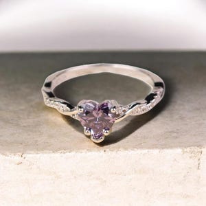 May include: A silver ring with a heart-shaped, pale purple gemstone. The band has a twisted design with small, clear accent stones. The ring is set against a neutral background, highlighting the jewellery's details.