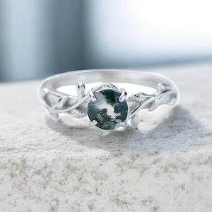 Puede incluir: Un anillo de plata con una piedra redonda de ágata musgosa. El anillo presenta un diseño de hojas en la banda y alrededor de la piedra. La piedra de ágata musgosa es una mezcla de verde y blanco, engastada en una banda de plata.