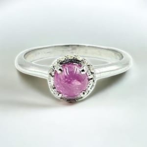 Może przedstawiać: Srebrny pierścionek z okrągłym, różowym kamieniem szlachetnym pośrodku. Kamień szlachetny jest osadzony w szczegółowej oprawie. Obręcz pierścionka jest gładka i zaokrąglona.