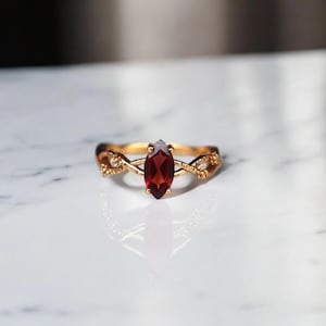 Könnte beinhalten: Ein goldener Ring mit einem marquiseförmigen, tiefroten Edelstein. Das Band weist ein kompliziertes, florales Design auf. Der Ring liegt auf einer weißen Marmoroberfläche, die das Licht reflektiert.