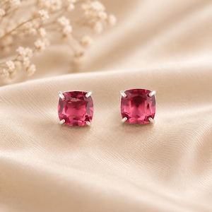 May include: A pair of stud earrings featuring cushion-cut, deep pinkish-red gemstones set in silver-toned metal prongs. Each earring displays a faceted surface that reflects light, showcasing a vibrant, saturated color and a classic square shape.