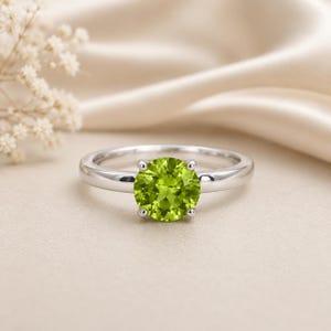 May include: A silver-toned ring featuring a round, faceted, lime-green gemstone set in a four-prong mounting. The band is smooth and polished, showcasing a simple design that highlights the vibrant color and clarity of the central stone.