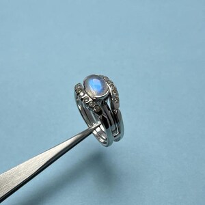 Könnte beinhalten: Ein silbernes Ring-Set mit einem ovalen Mondstein, der ein blau-weißes Schimmern aufweist. Das Band ist mit kleinen, klaren Edelsteinen verziert. Der Ring wird von einer Pinzette vor einem hellblauen Hintergrund gehalten.