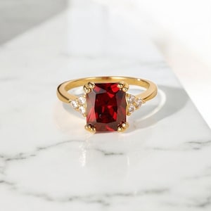 Puede incluir: Un anillo dorado con una gran piedra preciosa rectangular de color rojo intenso como pieza central. El anillo está adornado con piedras preciosas más pequeñas y claras a ambos lados de la piedra principal, sobre un fondo de mármol blanco.