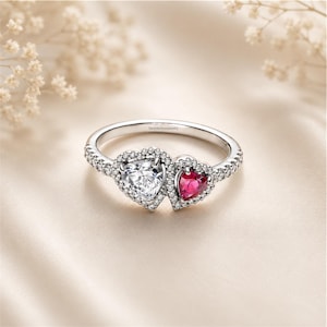 May include: A silver-toned ring featuring two heart-shaped stones, one clear and one deep red, both surrounded by small pavé-set stones. The band is also encrusted with small clear stones and includes the text SanjidaSilverjewelry engraved inside.