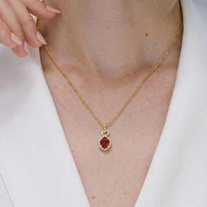 May include: A gold-colored necklace with a pendant featuring a large, oval-shaped red gemstone surrounded by small, clear stones. The pendant is suspended from a delicate gold chain. The necklace is worn against a white background.