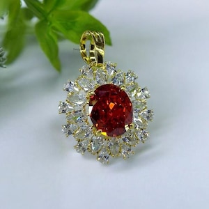 May include: A gold pendant with a large, oval-cut, deep red gemstone at the centre, encircled by smaller, clear, teardrop-shaped stones. The pendant has a gold bail for hanging.