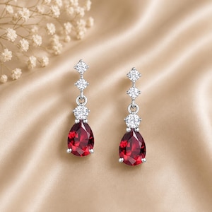 May include: A pair of silver-toned drop earrings featuring three small clear round stones arranged vertically above a large pear-shaped deep red stone. The earrings have a polished metallic finish and a secure setting for each of the faceted gemstones.