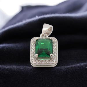 May include: A silver pendant with a rectangular, emerald-green gemstone at its center. The gemstone is surrounded by a halo of small, clear stones. The pendant has a simple, elegant design, suitable for everyday wear or special occasions.